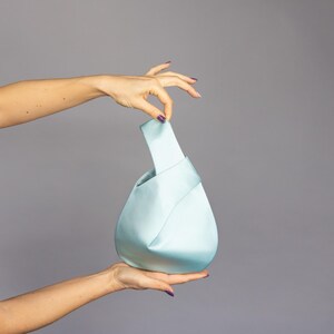 Könnte beinhalten: Hellblaue Satin-Handtasche mit geknotetem Henkel. Die Tasche hat eine abgerundete Form und wird von zwei Händen vor grauem Hintergrund gehalten. Der Griff der Tasche ist in einem helleren Blauton gehalten.