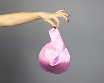 May include: A light pink satin bag with a knotted handle. The bag has a rounded shape and is held up by a hand against a gray background. The satin material has a slight sheen.
