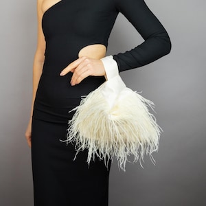 May include: A black, one-shoulder dress with a cut-out detail. A white satin clutch with cream-coloured feathers is held by the person. The dress has a long sleeve on the right arm.