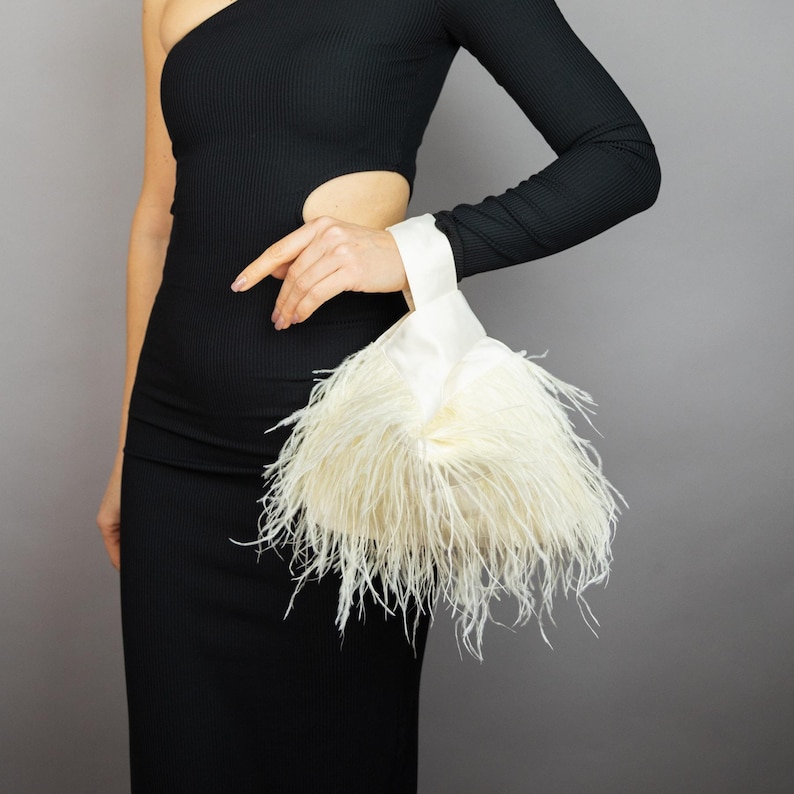 May include: A black, one-shoulder dress with a cut-out detail. A white satin clutch with cream-coloured feathers is held by the person. The dress has a long sleeve on the right arm.