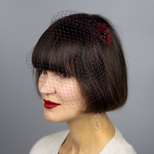 May include: A woman with short, dark brown hair wearing a burgundy veil with a burgundy flower accent. She is wearing red lipstick and a silver earring.
