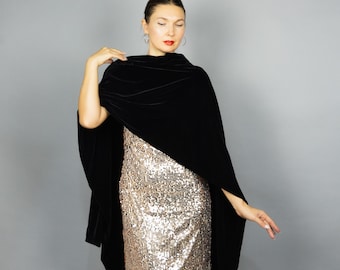 Puede incluir: Una mujer con una capa de terciopelo negro sobre un vestido de lentejuelas doradas. La capa cae sobre los hombros y los brazos, creando una silueta fluida. El vestido brilla con lentejuelas doradas, reflejando la luz. El fondo es gris liso.