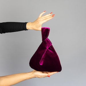 Pode incluir: Uma bolsa de mão de veludo magenta escuro com uma alça com nós. A bolsa em forma de triângulo é segurada por uma pessoa com esmalte vermelho em um fundo cinza. A textura macia e a cor rica da bolsa são destacadas.