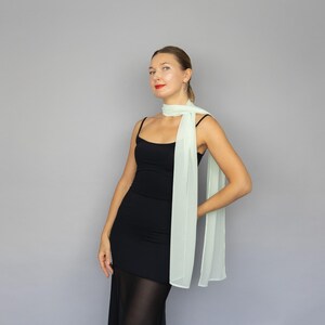 May include: A woman wearing a black dress and a light green scarf. The scarf is draped around the neck and hangs down the front. The background is a solid gray.