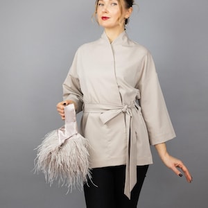 May include: A beige kimono jacket with a tie belt, paired with black trousers. The jacket features wide sleeves. A light pink satin clutch, adorned with fluffy feathers, is held in the hand.