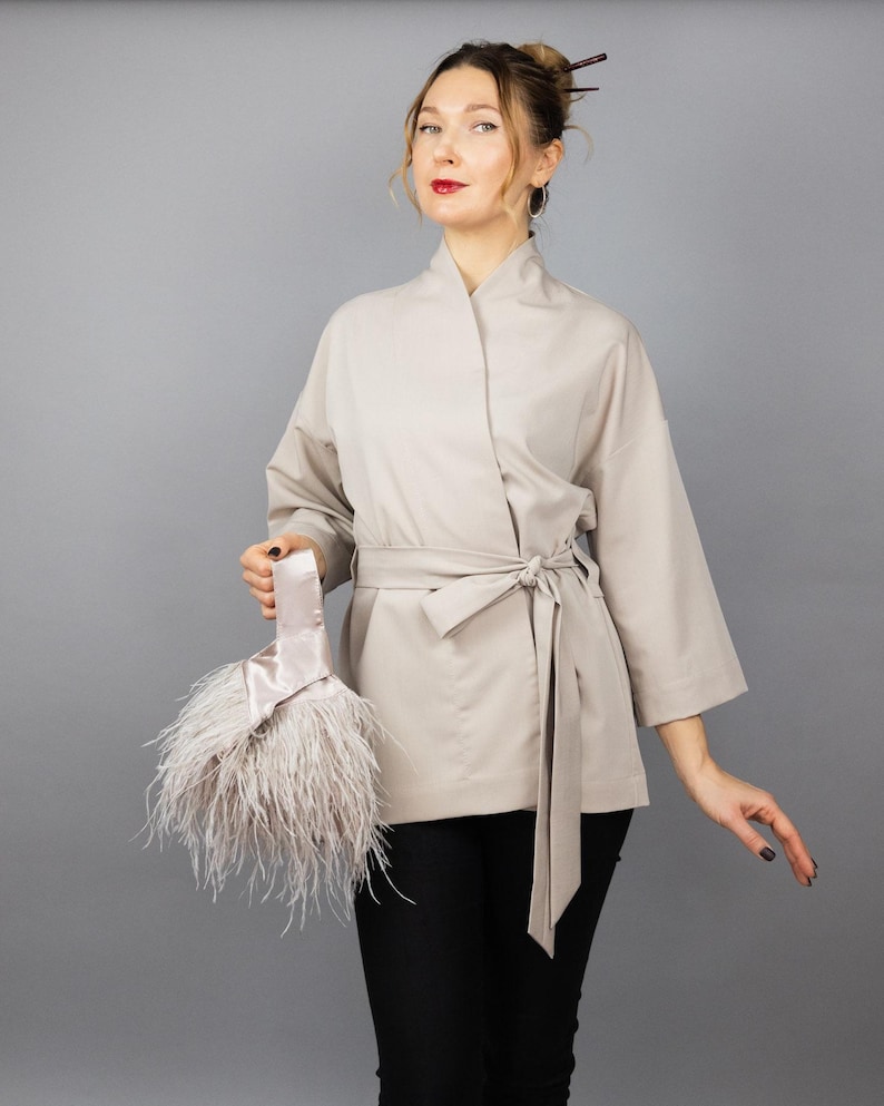 May include: A beige kimono jacket with a tie belt, paired with black trousers. The jacket features wide sleeves. A light pink satin clutch, adorned with fluffy feathers, is held in the hand.