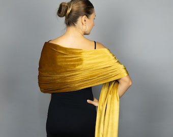 May include: Mustard yellow velvet shawl draped over a black dress. The shawl is wrapped around the shoulders and arms, with one end hanging down. The fabric has a soft, luxurious appearance, and the color is rich and vibrant.