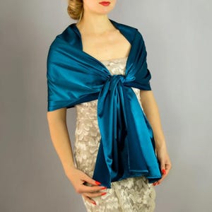 May include: A teal blue satin shawl draped over a person's shoulders and tied at the front. The shawl is soft and flowing, and would be a beautiful addition to any outfit.