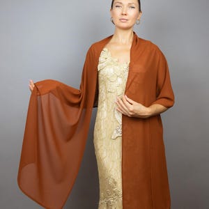 May include: A rust-colored chiffon shawl draped over a person wearing a gold lace dress. The shawl is sheer and lightweight, with a flowing drape.