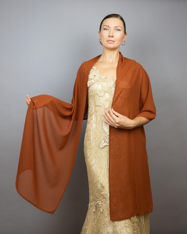 May include: A rust-colored chiffon shawl draped over a person wearing a gold lace dress. The shawl is sheer and lightweight, with a flowing drape.