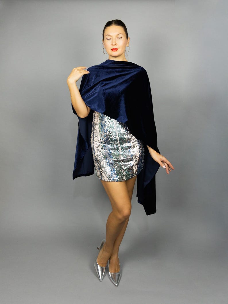 May include: A navy blue velvet cape draped over a silver sequined mini dress. The cape has a soft, flowing texture. Silver pointed-toe heels complete the look, creating an elegant ensemble.