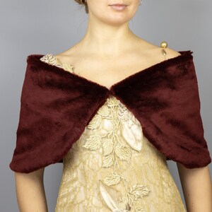 K&ouml;nnte beinhalten: Ein burgunderfarbener Kunstfellschal mit V-Ausschnitt. Der Schal ist &uuml;ber ein goldenes Kleid mit Blumenstickerei gelegt.
