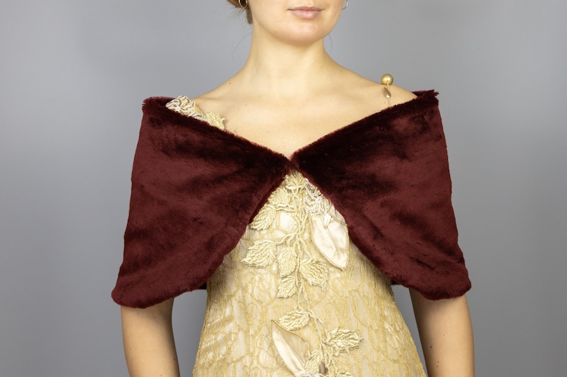 K&ouml;nnte beinhalten: Ein burgunderfarbener Kunstfellschal mit V-Ausschnitt. Der Schal ist &uuml;ber ein goldenes Kleid mit Blumenstickerei gelegt.