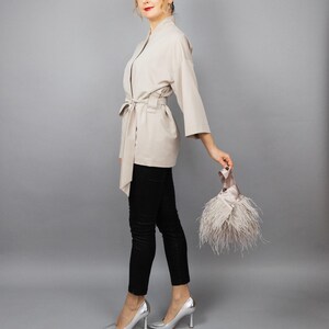 May include: A beige kimono jacket with a tie belt, worn with black trousers and silver heels. A small clutch bag with feathers is held in the hand. The background is a plain grey.