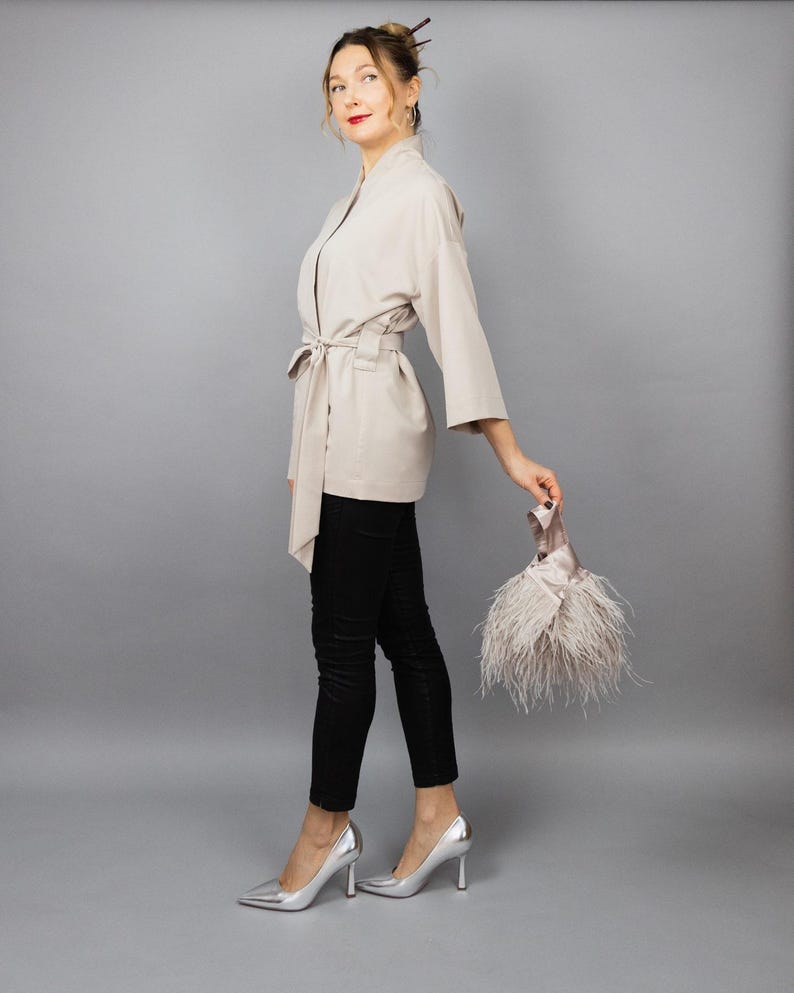 May include: A beige kimono jacket with a tie belt, worn with black trousers and silver heels. A small clutch bag with feathers is held in the hand. The background is a plain grey.