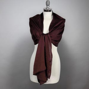 May include: A burgundy satin shawl draped over a white mannequin. The shawl is tied in a knot at the front and the ends are left to hang down.