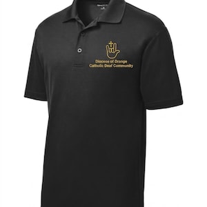 Puede incluir: Polo negro con cuello y mangas cortas. La camisa presenta un logotipo dorado y el texto "Diocese of Orange Catholic Deaf Community". La camisa tiene una tapeta de tres botones.