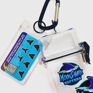 May include: A clear plastic container with a black carabiner clip and an ID tag. The ID tag has a blue background with shark tooth illustrations and text. The container has a "MoonShark Sifters" logo with a blue shark fin.