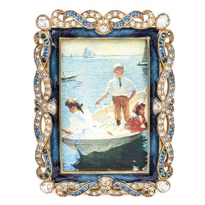 May include: A rectangular picture frame featuring a dark blue enamel border with a textured finish. The frame is adorned with gold-tone metal scrollwork, clear crystals, and blue rhinestones. It contains a print of people in a rowboat on water.