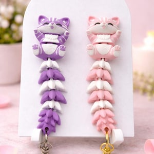 Puede incluir: Un par de pendientes con forma de gato. Un pendiente es morado con detalles blancos y un diseño floral colgante a juego. El otro pendiente es rosa con detalles blancos y un diseño floral colgante a juego. Ambos pendientes tienen cierres dorados.