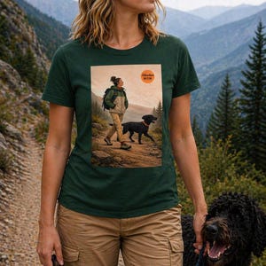 Peut inclure: T-shirt vert foncé avec un motif d'une personne faisant de la randonnée avec un chien, avec le texte "Adventure Mom" dans un cercle. La personne porte un pantalon kaki et le chien est noir. L'arrière-plan montre une chaîne de montagnes.