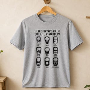 May include: A heather gray short-sleeve t-shirt featuring a black graphic print titled DETECTORIST'S FIELD GUIDE TO RING PULLS. The design displays nine illustrations of various metal ring pull tabs, each labeled with a specific year from 1963 to 2000-current.