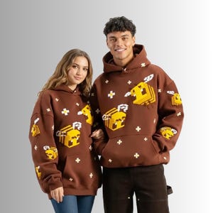 May include: Two brown hooded sweatshirts featuring a repeating pattern of yellow pixelated bees and small white crosses. Each hoodie has a front kangaroo pocket, long sleeves, and a drawstring hood, showcasing a consistent blocky graphic design.