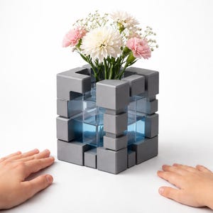 Könnte beinhalten: Eine graue, würfelförmige Vase mit einem transparenten, blau getönten Innenraum, die einen Strauß weißer und rosa Blumen enthält. Die Vase hat ein geometrisches Design, wobei die Würfelform aus kleineren, quadratischen Blöcken besteht.