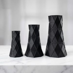 Puede incluir: Tres jarrones geométricos negros de diferentes alturas. Cada jarrón presenta un patrón triangular repetido, creando una superficie texturizada. Los jarrones se exhiben sobre una superficie de mármol blanco, ofreciendo una estética moderna y minimalista.