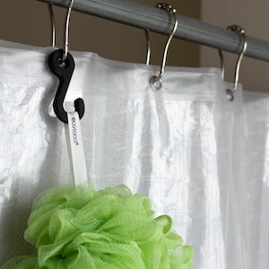 May include: A black shower curtain hook with a white tag that reads "ecotools" is attached to a shower curtain rod. A green mesh bath sponge hangs from the hook. The shower curtain is white and translucent.