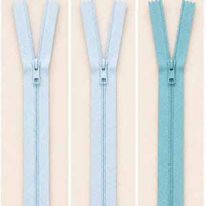 Puede incluir: Tres cremalleras abiertas en diferentes tonos de azul: azul cielo, azul polvo y azul turquesa. Cada cremallera está centrada sobre un fondo beige claro. El texto "SKY BLUE", "POWDER BLUE" y "TEAL BLUE" está impreso encima de cada cremallera.