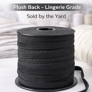 Op de afbeelding: Een spoel zwart 1,27 cm breed bh-elastiek met een zachte achterkant, geschikt voor lingerie. De tekst "Plush Back - Lingerie Grade" en "Sold by the Yard" zijn zichtbaar. Het elastiek is netjes op een zwarte spoel gewikkeld.