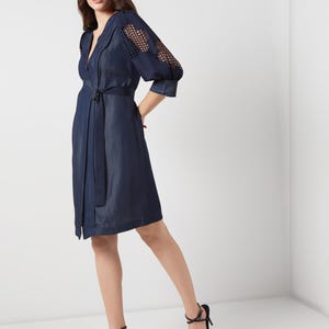 May include: A navy blue wrap dress with a notched collar and a matching belt. The dress features openwork detailing on the sleeves and is paired with navy blue high-heeled sandals. The dress is knee-length.