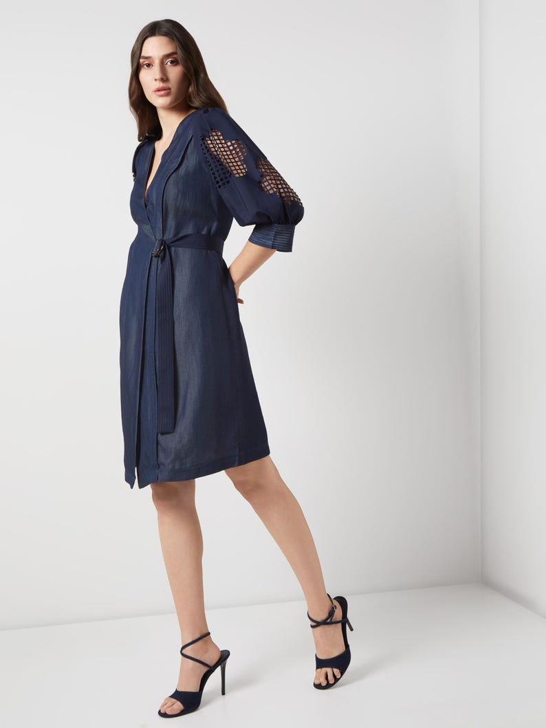 May include: A navy blue wrap dress with a notched collar and a matching belt. The dress features openwork detailing on the sleeves and is paired with navy blue high-heeled sandals. The dress is knee-length.