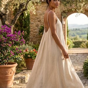 May include: A floor-length, sleeveless beige dress with a loose, flowing silhouette, thin shoulder straps, and a low-cut back design. The garment features a simple, unadorned aesthetic with a soft, textured appearance and a full, flared skirt.