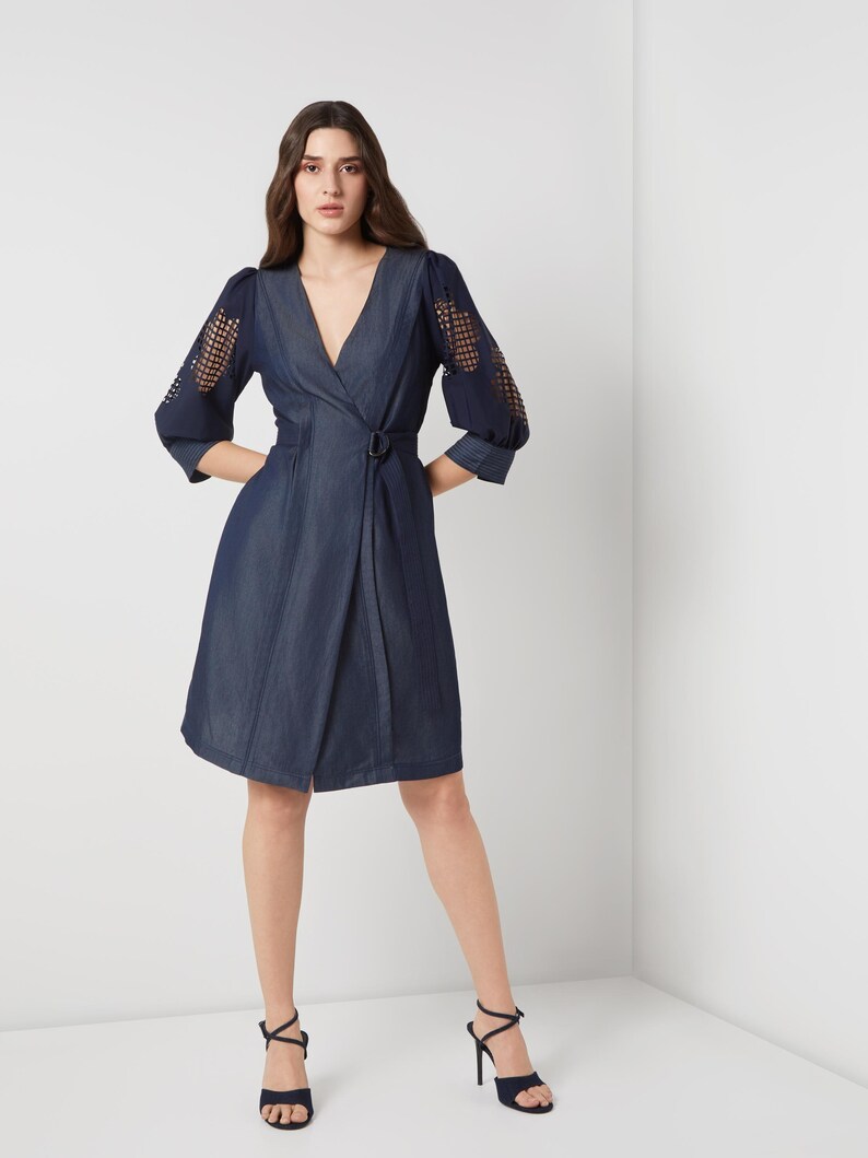 May include: A dark blue wrap dress with a V-neckline and a belt. The dress features sheer, open-weave sleeves and a knee-length skirt. The model is wearing black high-heeled sandals.