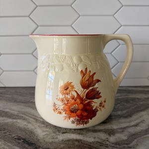 May include: Cream-colored ceramic pitcher with a red rim and a curved handle. The pitcher is decorated with a floral design in shades of orange, red, and brown. A grape vine design is embossed on the pitcher.