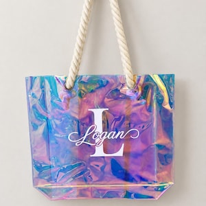 May include: A rectangular tote bag made of iridescent, semi-transparent material that reflects shades of blue, pink, and purple. The bag features thick, twisted white rope handles and a large white letter L with the name Logan printed over it.