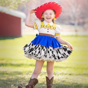 May include: A young girl dressed as a cowgirl, wearing a red cowboy hat, a white shirt with a yellow sheriff's star, a blue skirt with a black and white cow print, and brown cowboy boots. She is smiling and holding her hat.