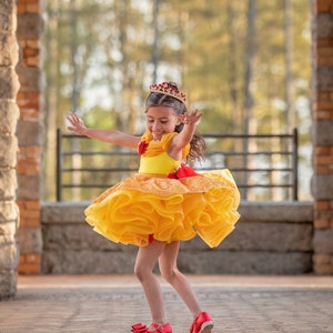 May include: A young girl in a yellow and orange ruffled dress and a red jeweled crown. She is wearing red shoes with bows and has her arms outstretched. The dress has a red bow at the waist.