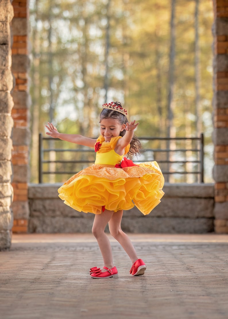 May include: A young girl in a yellow and orange ruffled dress and a red jeweled crown. She is wearing red shoes with bows and has her arms outstretched. The dress has a red bow at the waist.