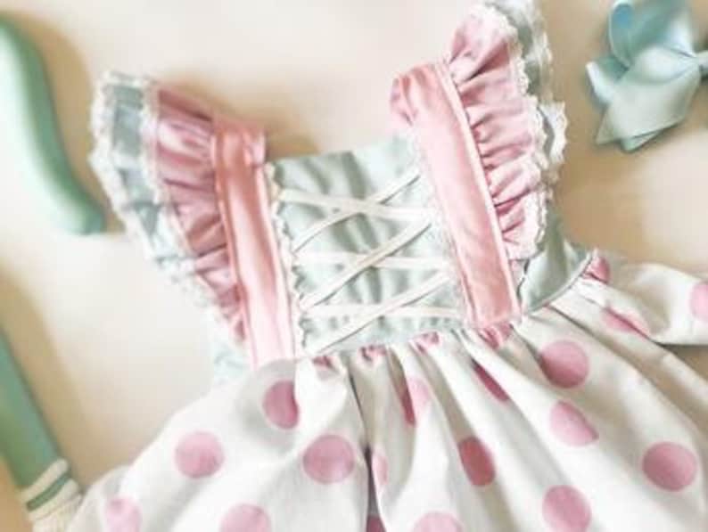 May include: A light blue and pink polka dot dress with white lace trim and pink ruffles on the straps. The dress has a white criss-cross back.