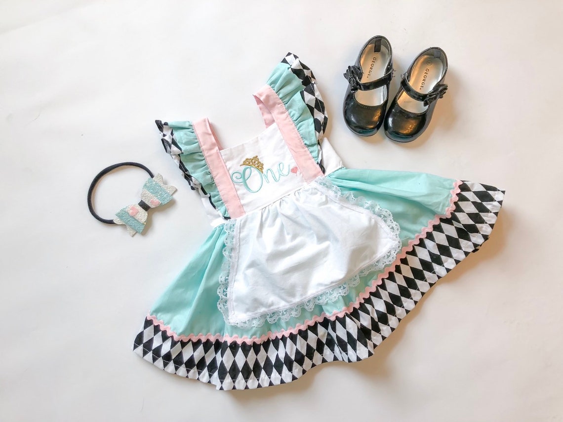 May include: A light blue and white dress with a black and white diamond pattern trim. The dress has a white apron with lace trim and a pink and white ruffle trim. The dress has the word 'One' embroidered on the front. The dress is paired with a black headband with a silver and white bow. The dress is also paired with two black shoes with a bow on the toe.