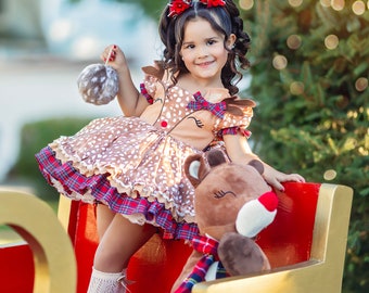 May include: A young girl wearing a brown and red reindeer dress with a plaid skirt sits on a red sleigh with a brown teddy bear. She is holding a white and brown ornament. The sleigh is decorated with gold trim.