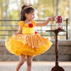 May include: A young girl in a yellow dress with a red rose detail and a crown, pointing at a glass cloche with a red rose inside. The dress has a layered skirt and a red bow. Red shoes complete the outfit. A dark wood table holds the cloche.