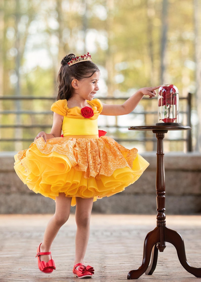 May include: A young girl in a yellow dress with a red rose detail and a crown, pointing at a glass cloche with a red rose inside. The dress has a layered skirt and a red bow. Red shoes complete the outfit. A dark wood table holds the cloche.