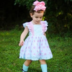 May include: A light blue and white polka dot dress with pink and white ruffled straps and a pink bow headband. The dress has a white lace overlay and a ruffled hem.