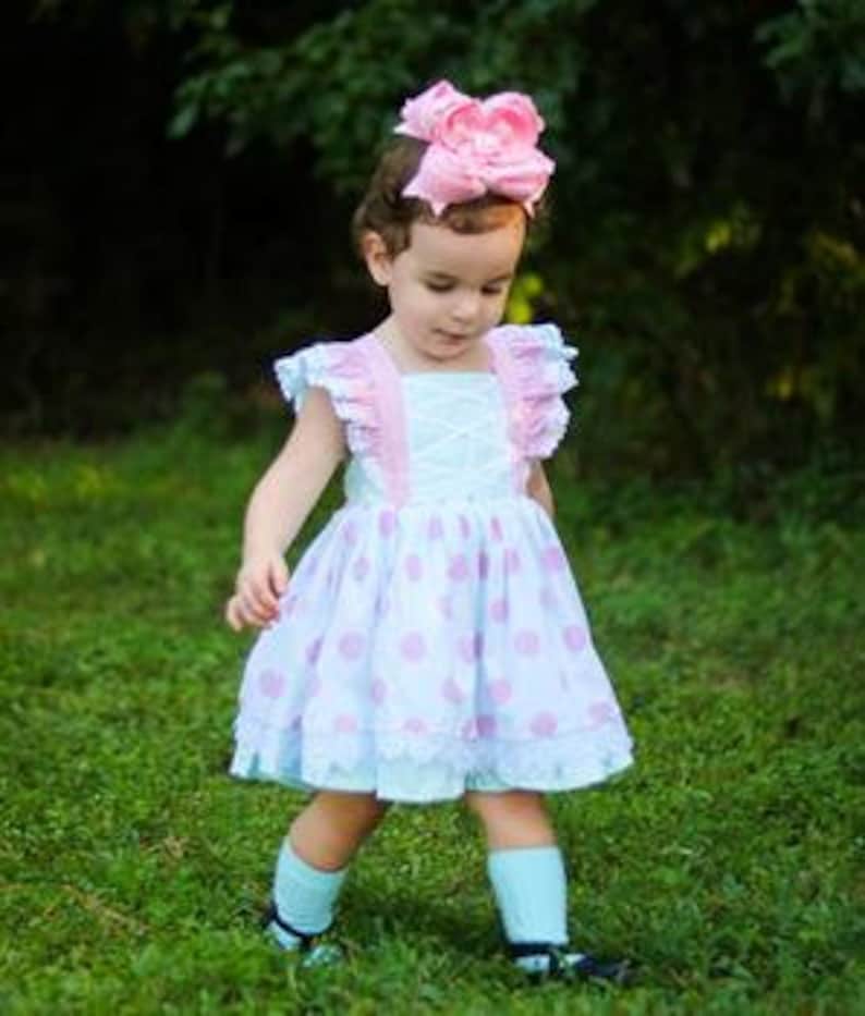 May include: A light blue and white polka dot dress with pink and white ruffled straps and a pink bow headband. The dress has a white lace overlay and a ruffled hem.