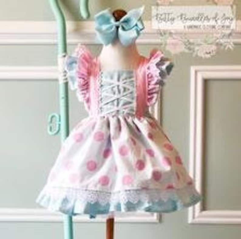 May include: A blue and pink polka dot dress with a white lace trim and pink ruffle sleeves. The dress has a criss-cross bodice and is styled with a blue bow.