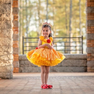 May include: A young girl wearing a yellow and orange dress with a red rose and a crown. The dress has a layered skirt and ruffled sleeves. She is wearing red shoes and standing in front of a stone archway.